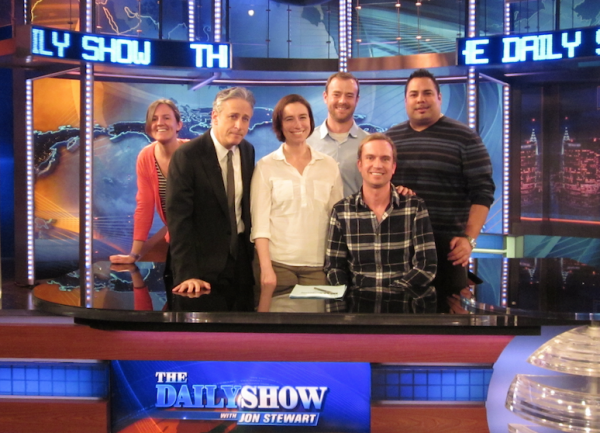 Jon Stewart with 5 of the veterans posing behind the Daily Show desk