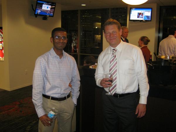 2 men pose for photo- 1 holding a bottle of water, 1 a glass of wine