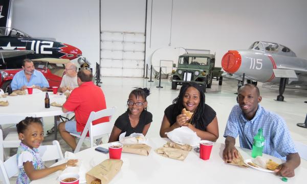 Family sitting at round table eating, jeep and plane in background