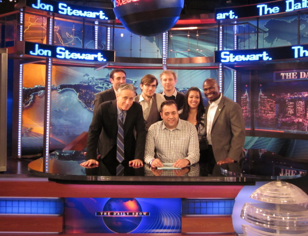 Jon Stewart with 6 of the veterans posing behind the Daily Show desk